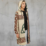 A woman wearing a hooded longline cardigan with a beige base and red, green, and black Celtic knot patterns, styled over a black top and white pants against a light gray studio background.