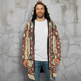 A man wearing a long hooded cardigan with a beige base and a bold red, black, and green Celtic knot pattern, layered over a white t-shirt and blue jeans, against a plain white background.