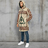 A man wearing a hooded long-sleeve tunic top with a beige base and red and green Celtic knot patterns, paired with blue jeans and white sneakers, against a plain white background.