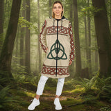 A woman modeling a beige hooded tunic dress with a green Celtic triquetra symbol on the front and red-brown Celtic knot patterns on the sleeves and hem, styled with white pants and white sneakers against a white studio background.