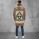 A man viewed from behind wearing a long hooded jacket with a beige base, red and brown Celtic knot borders, and a large green triquetra symbol on the back, paired with blue jeans and white sneakers against a white studio background.