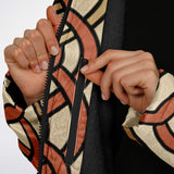 Close-up of a person's hands unzipping a patterned jacket with a black lining. The jacket features a geometric design in beige, black, and terracotta orange.