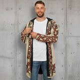 A man wearing a beige open-front cardigan with a bold black and rust-orange geometric pattern, layered over a white t-shirt and blue jeans, against a plain white studio background.