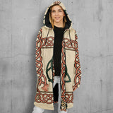 A woman wearing a hooded longline coat with an all-over Celtic knot pattern in beige, terracotta, and green, styled over a black top and light gray pants against a plain white background.